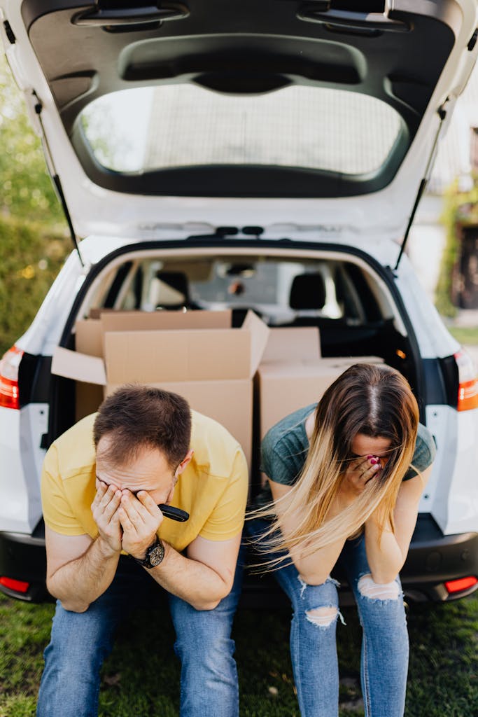 A distressed couple sits by a car full of boxes, symbolizing moving stress.